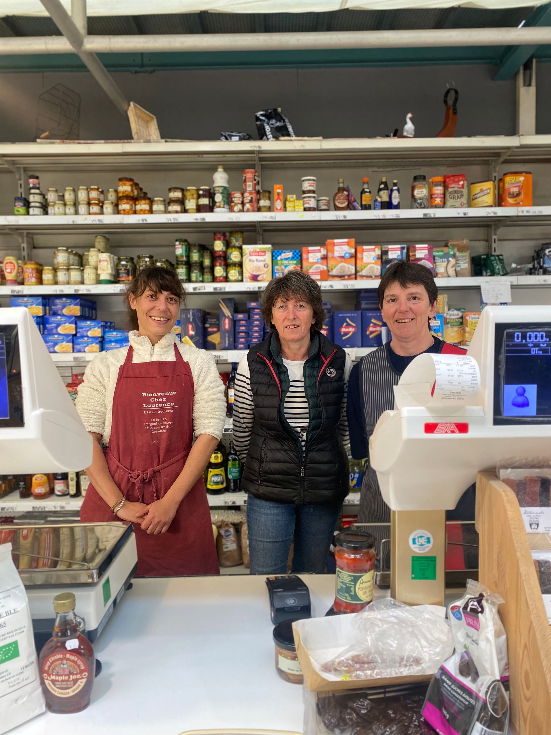 Trois employés de magasin sourient derrière une caisse, dans un magasin dont on aperçoit les rayons d'épicerie en arrière-plan.