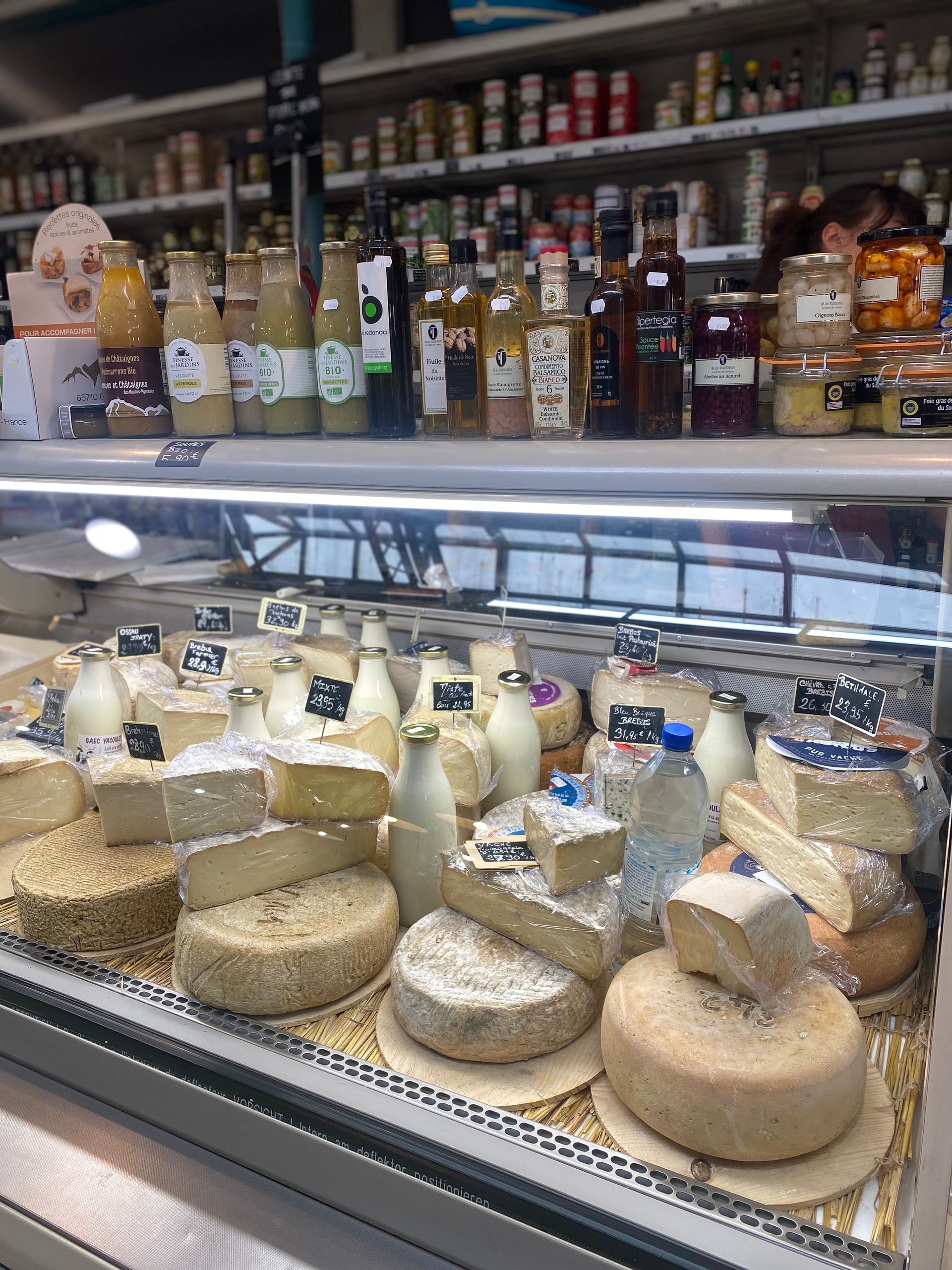 Une vitrine de marché remplie de meules et de portions de fromages artisanaux de toutes sortes, avec des produits en bouteille sur les étagères au-dessus.