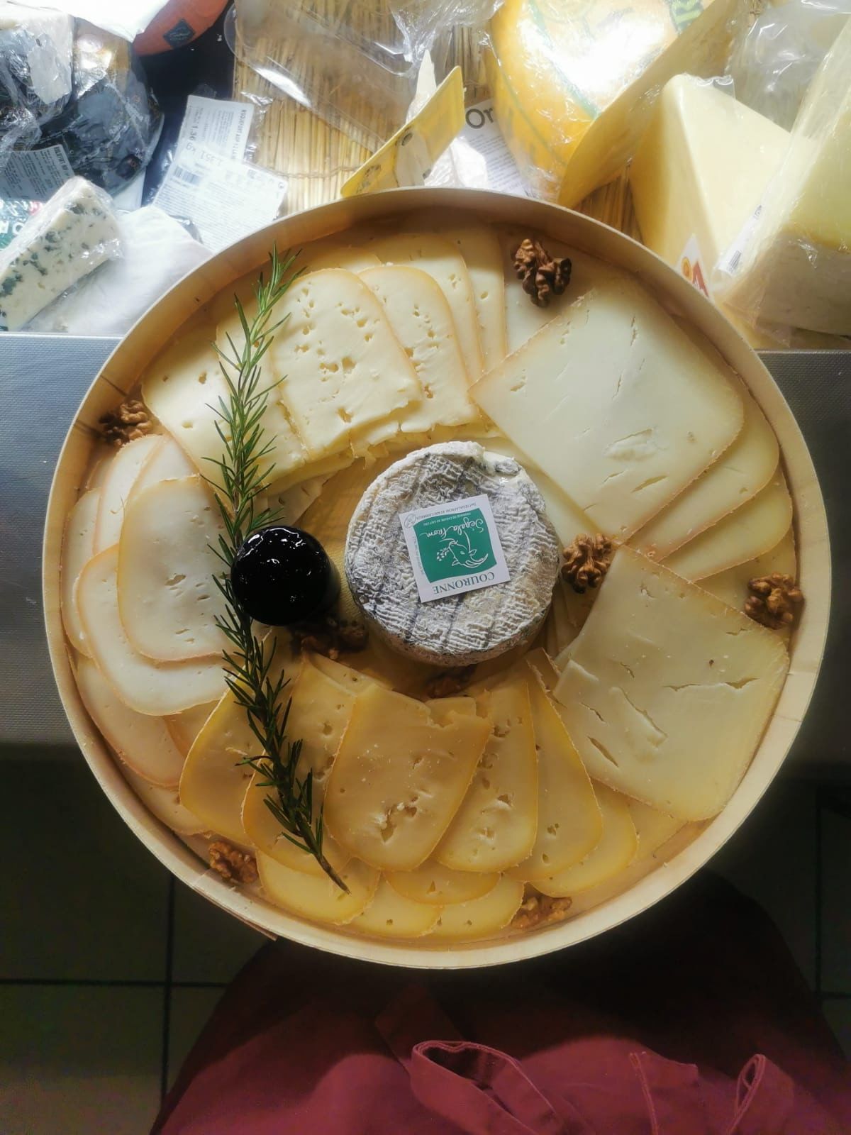 Un plateau rond en bois contient divers fromages tranchés, un petit fromage entier avec une étiquette, un brin de romarin et des noix.