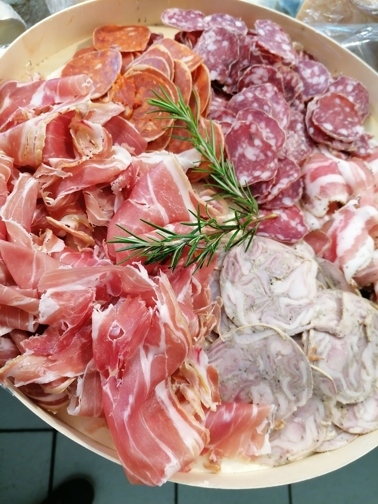Un plateau en bois garni de charcuteries assorties, notamment du prosciutto tranché, du salami et du fromage de tête, le tout parsemé de romarin.