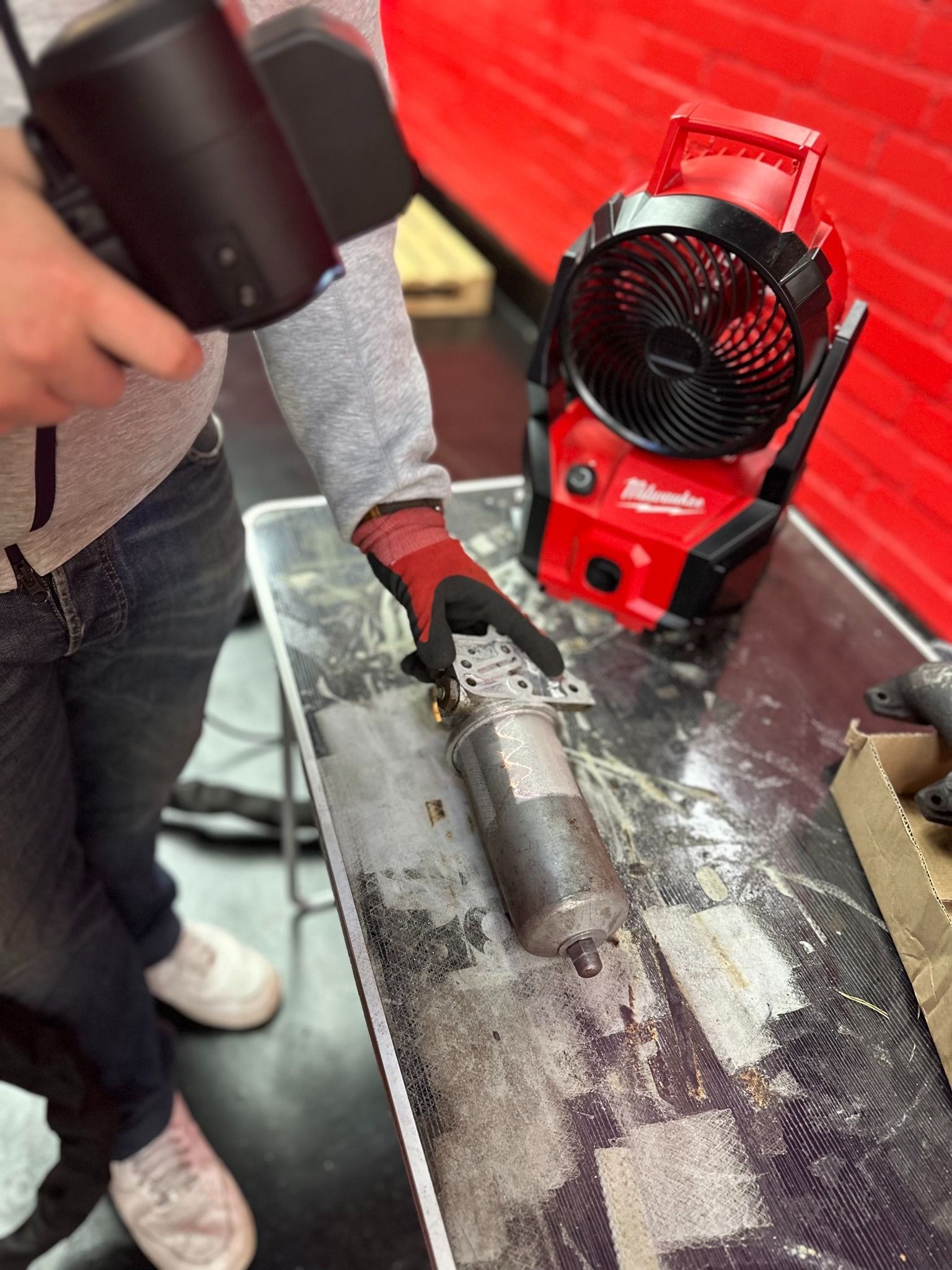 Skanneria pitelevä henkilö osoittaa pöydällä olevia metallisylintereitä, taustalla Milwaukee-tuuletin.