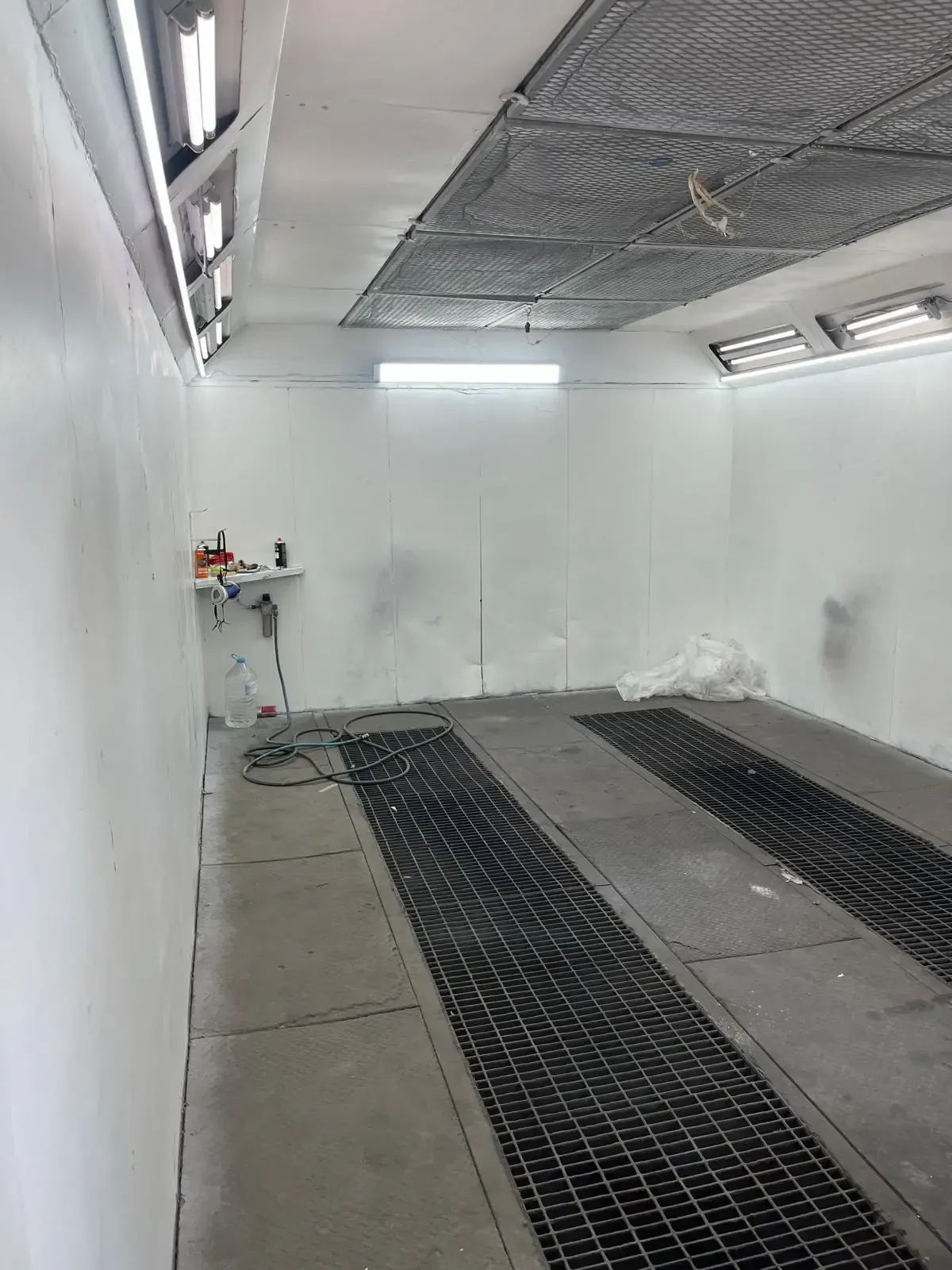 Vista interior de una cabina de pintura en aerosol, con una rejilla de piso, luces superiores y una estación de trabajo.