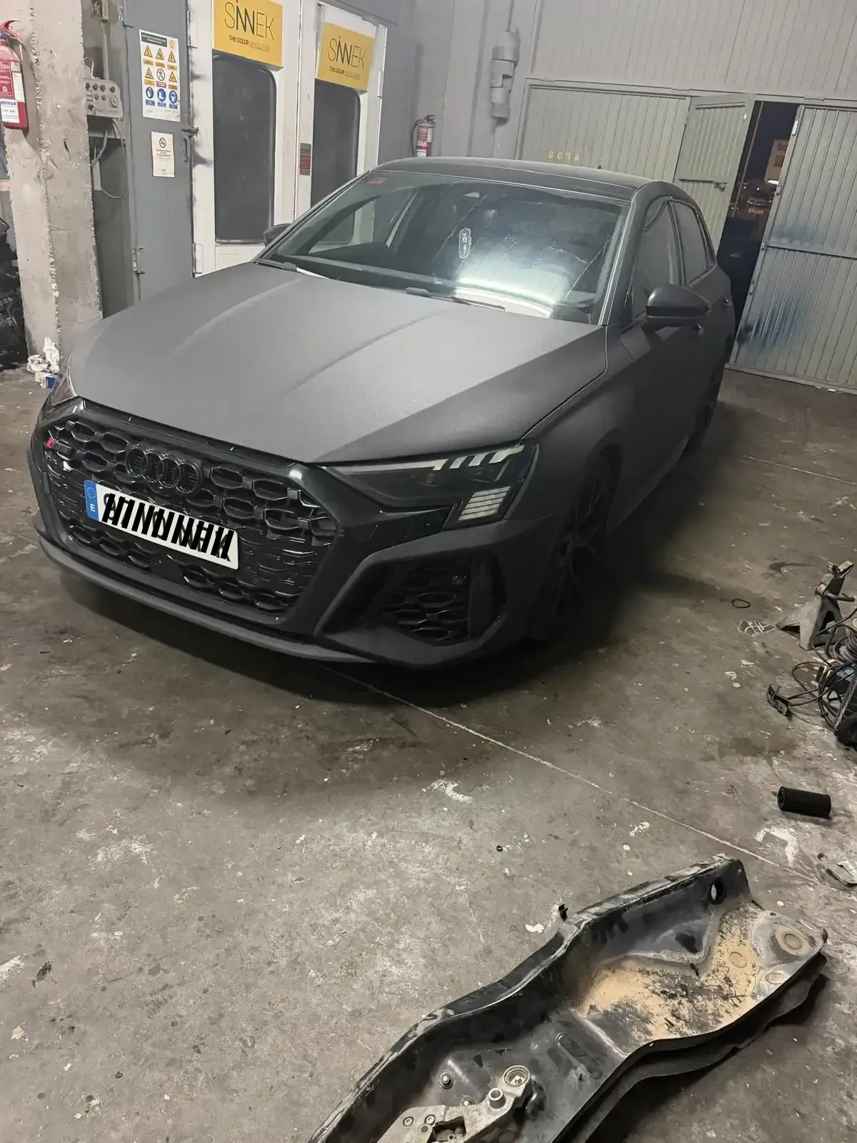 Coche negro en un elevador en un garaje con herramientas y equipos al fondo.