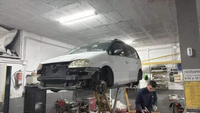 Coche subido en un elevador en un taller de reparación; mecánico trabajando debajo; falta el parachoques delantero del coche.
