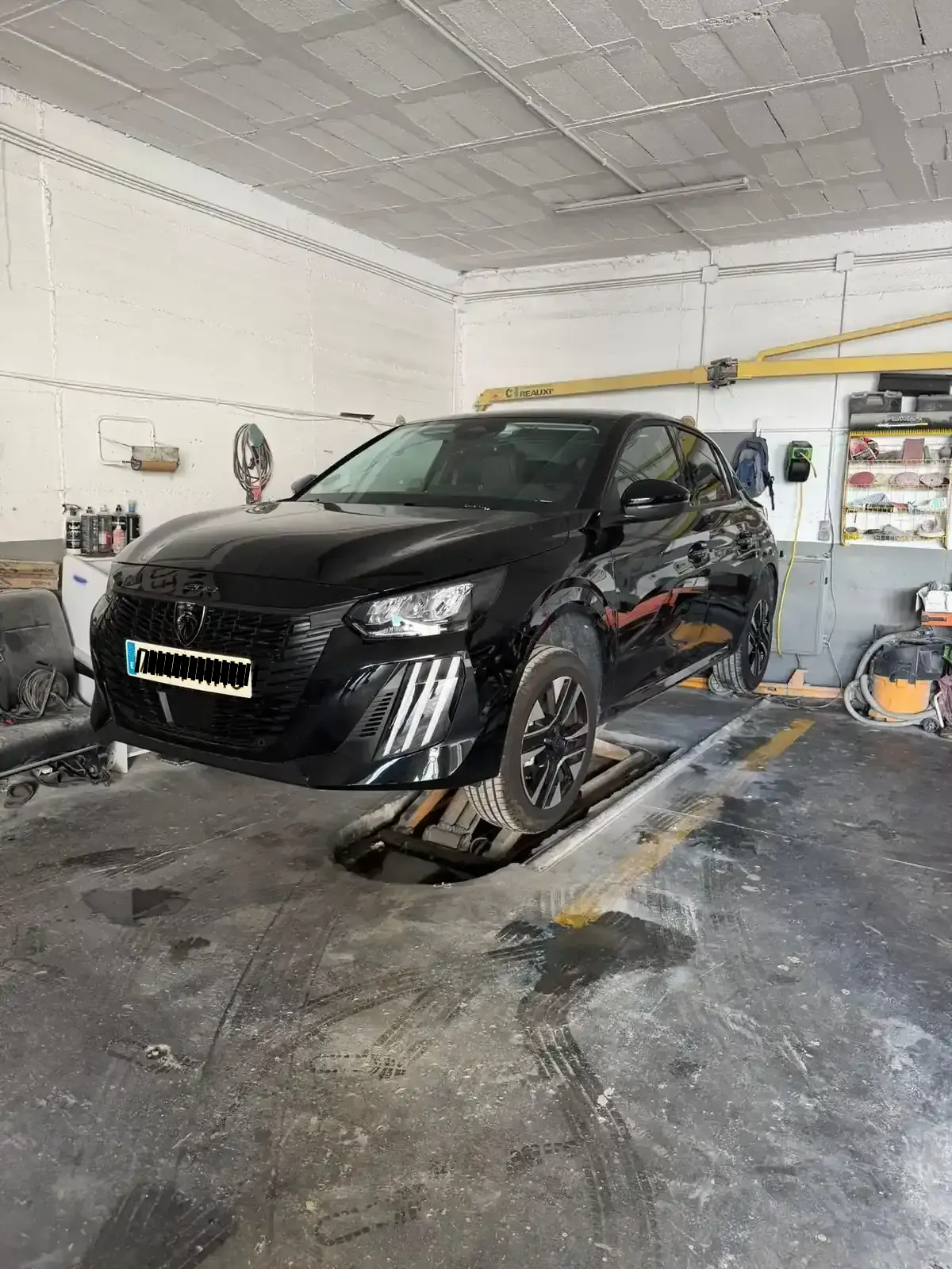 Coche negro en un elevador en un garaje con herramientas y equipos al fondo.