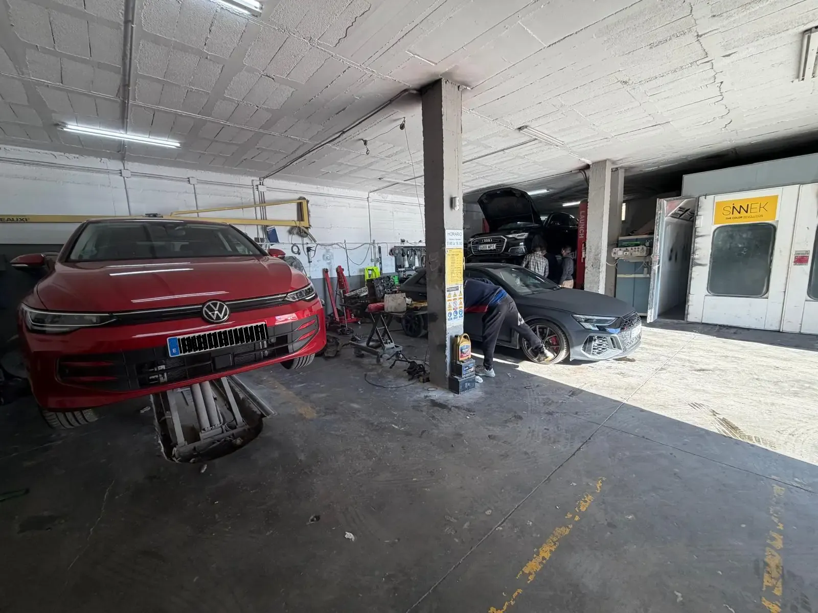Coche rojo en un elevador en un taller de reparación de automóviles; mecánico trabajando en otro coche.