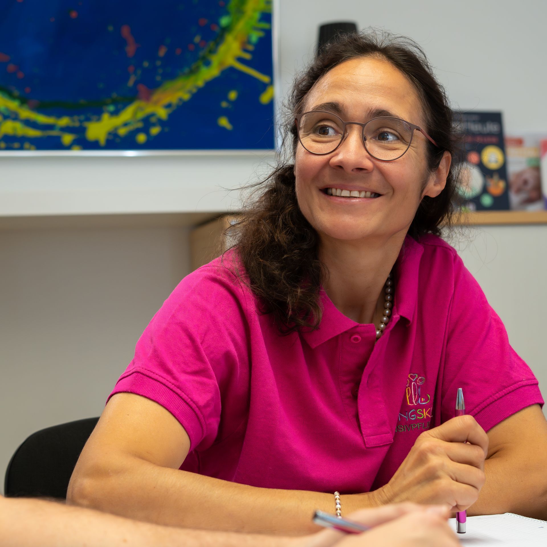 Kleinkind spielt mit einer Spielzeugeisenbahn und Bauklötzen an einem Tisch; im Hintergrund sind eine Betreuungsperson 