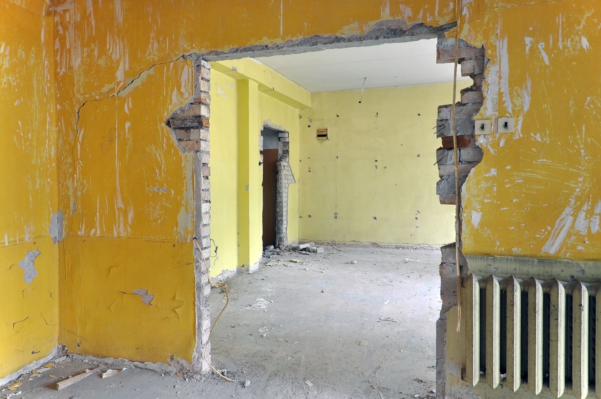 Une pièce jaune avec une grande porte en mauvais état donnant sur une pièce adjacente aux murs jaune clair et équipée d'un radiateur.