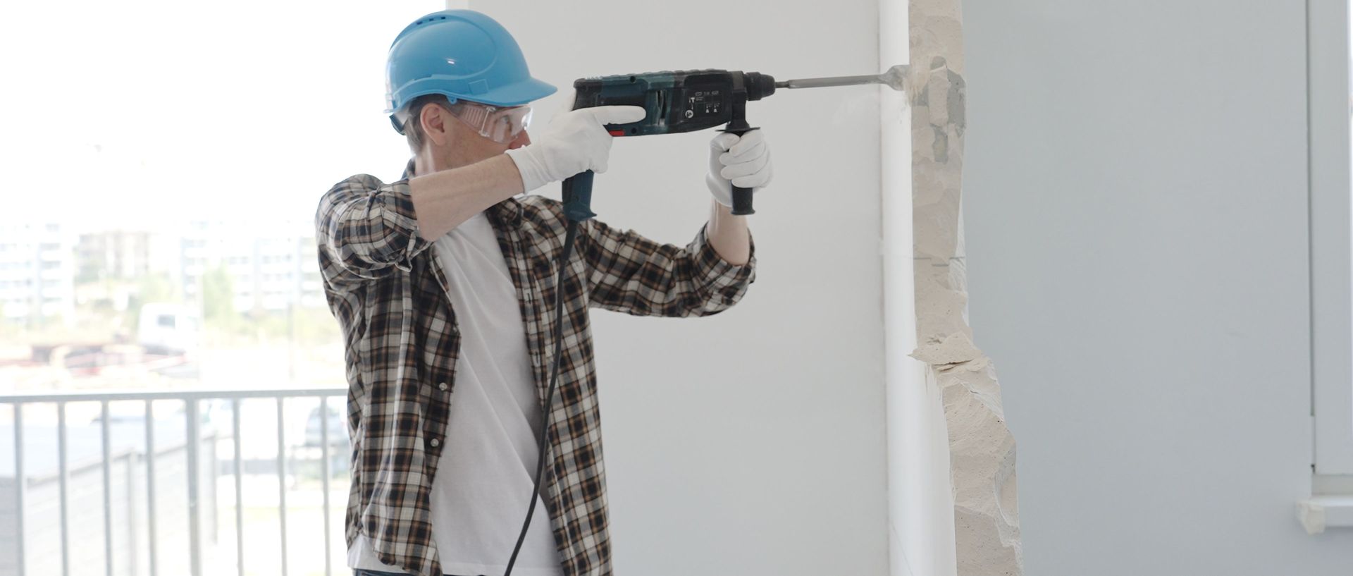 Dans un intérieur moderne et lumineux, une personne portant un casque de chantier et une chemise à carreaux utilise une perceuse électrique sur un mur.