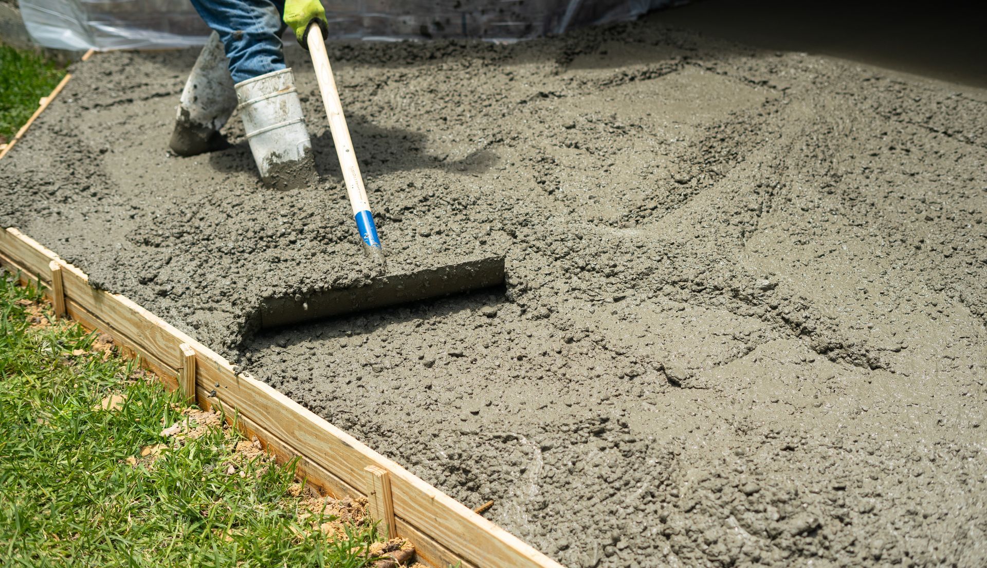Un ouvrier en bottes de travail utilise un râteau à béton pour étaler du béton gris et humide à l'intérieur d'un cadre en bois sur une pelouse.