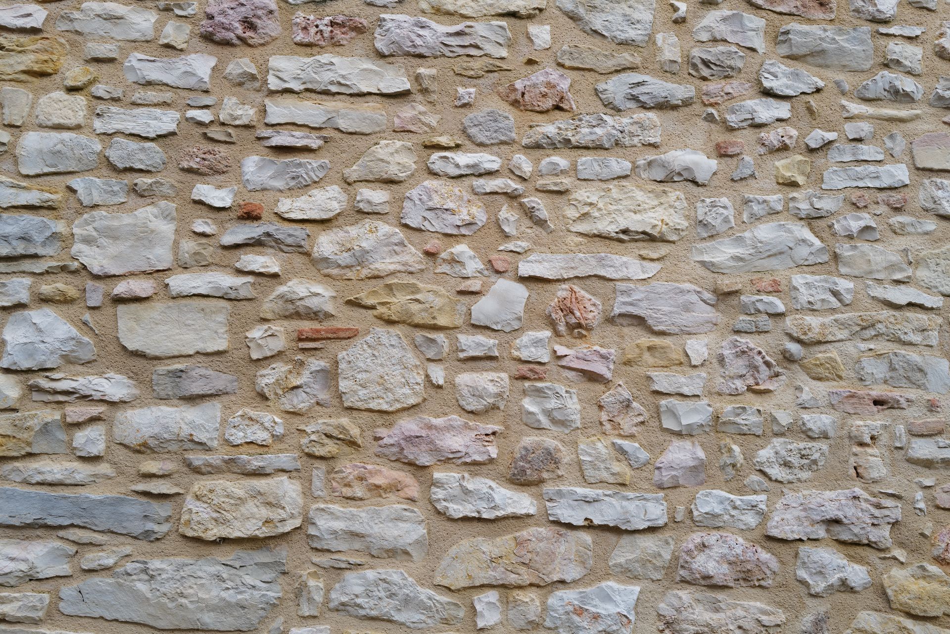 Un mur texturé composé de pierres des champs rugueuses et irrégulières, gris clair et beige, posées dans un mortier épais et clair.