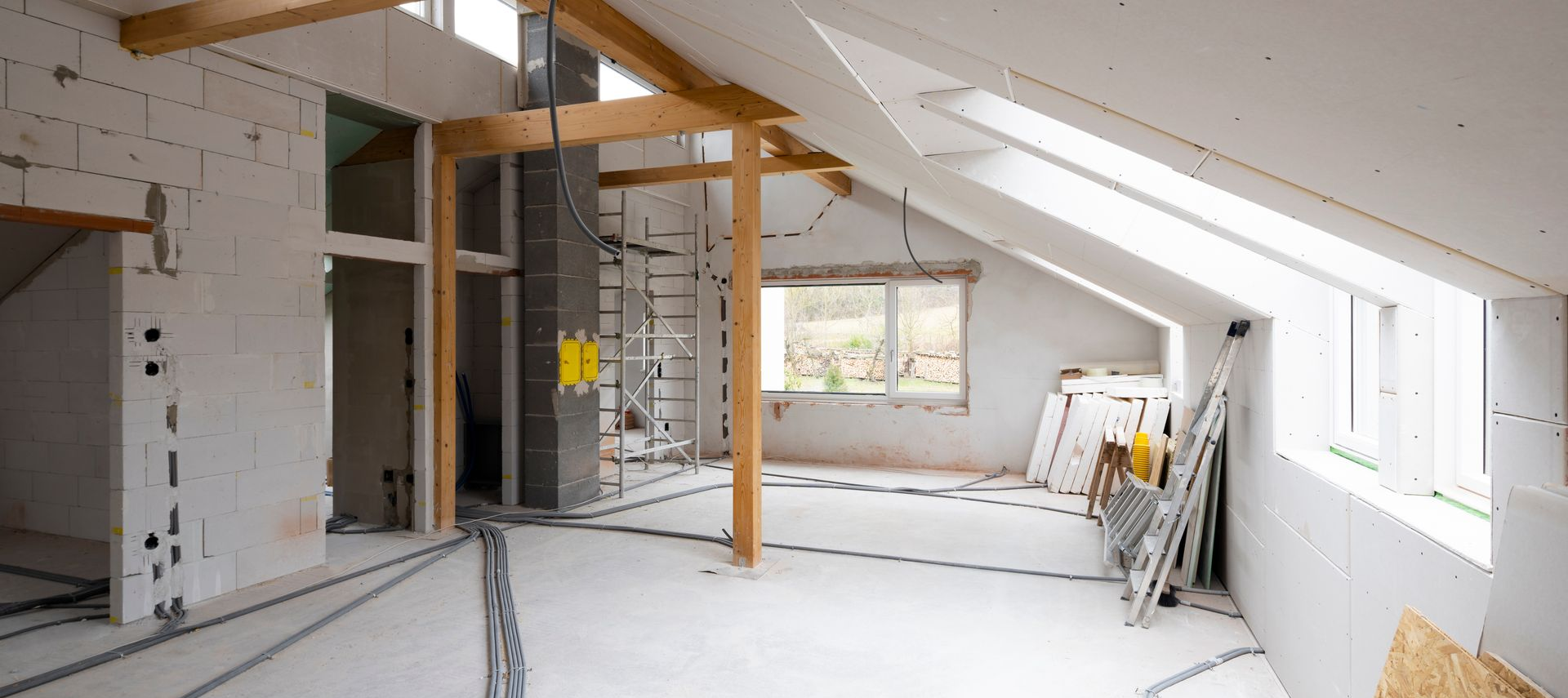 Une pièce mansardée inachevée en cours d'aménagement, avec des poutres en bois apparentes, des murs en plaques de plâtre et des puits de lumière.
