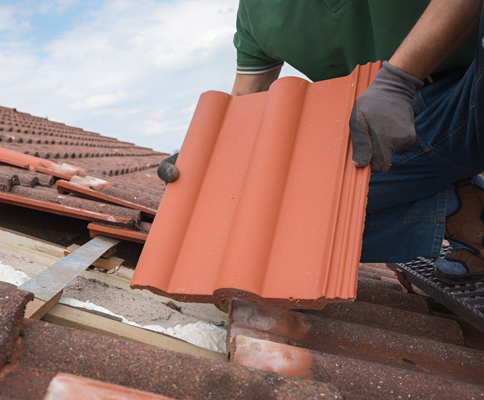 Un ouvrier portant des gants installe une nouvelle tuile rouge en terre cuite sur un toit résidentiel en pente.