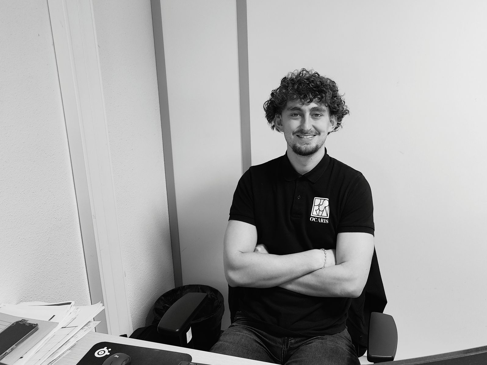 Maxence Gantzer, bras croisés, souriant dans un bureau, vêtu d'un polo noir.