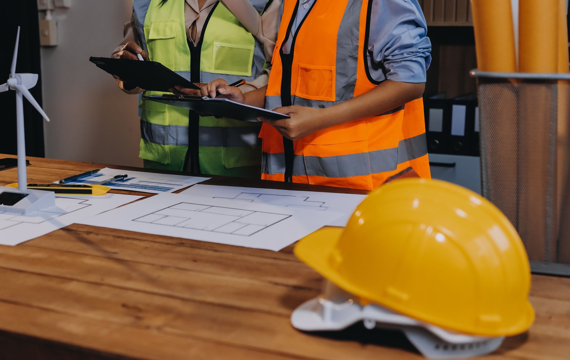 Deux personnes portant des gilets de sécurité examinent des plans sur un bureau avec un casque de sécurité jaune.