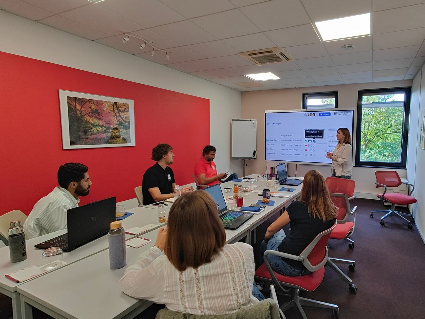 Une équipe de personnes lors d'une réunion, avec une femme présentant devant un écran dans une pièce.