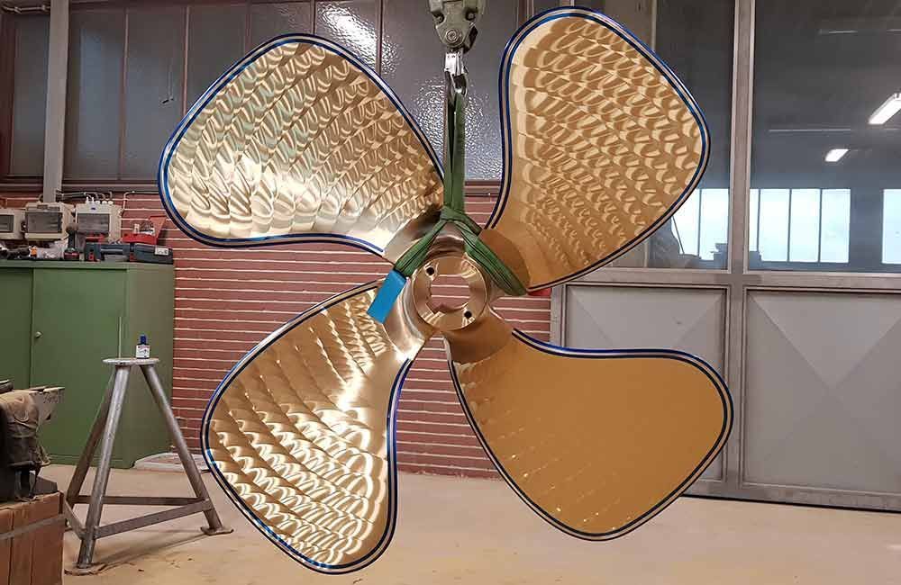 Ein großer, goldfarbener Propeller mit drei Blättern hängt an einem Hebezeug in einer Werkstatt.