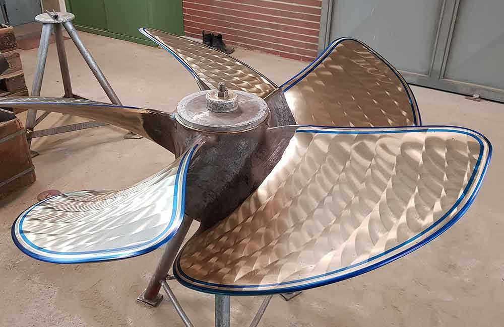 Großer, bronzener Schiffspropeller mit fünf Blättern, montiert auf einem Ständer. Die Blätter weisen silberne und goldene Verzierungen mit einem blauen Rand auf.