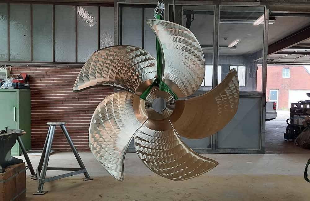 In einer Werkstatt hängt eine große, glänzende Schiffsschraube an grünen Gurten.