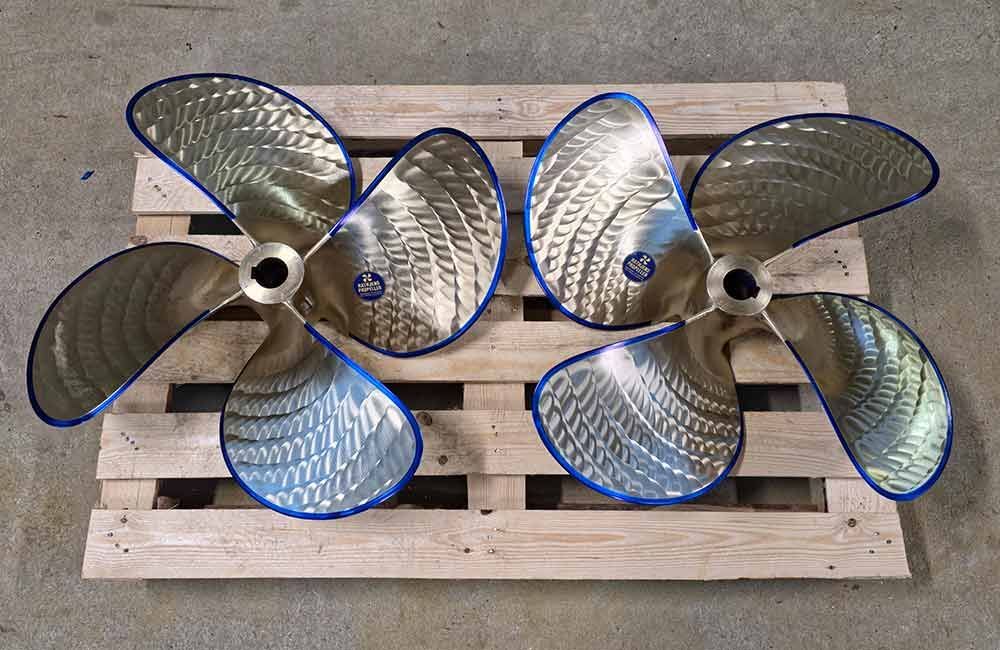 Zwei glänzende, vierblättrige Bootsschrauben mit blauem Rand, die auf einer Holzpalette ruhen.