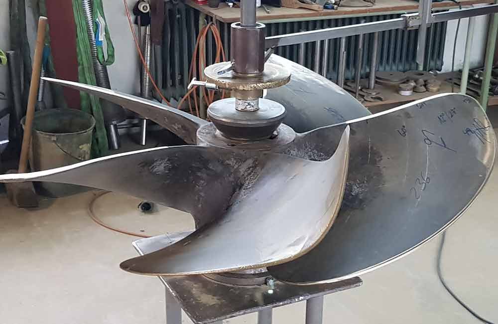 Großer Metallpropeller mit gebogenen Blättern, montiert auf einem Ständer, wahrscheinlich für ein Schiff.