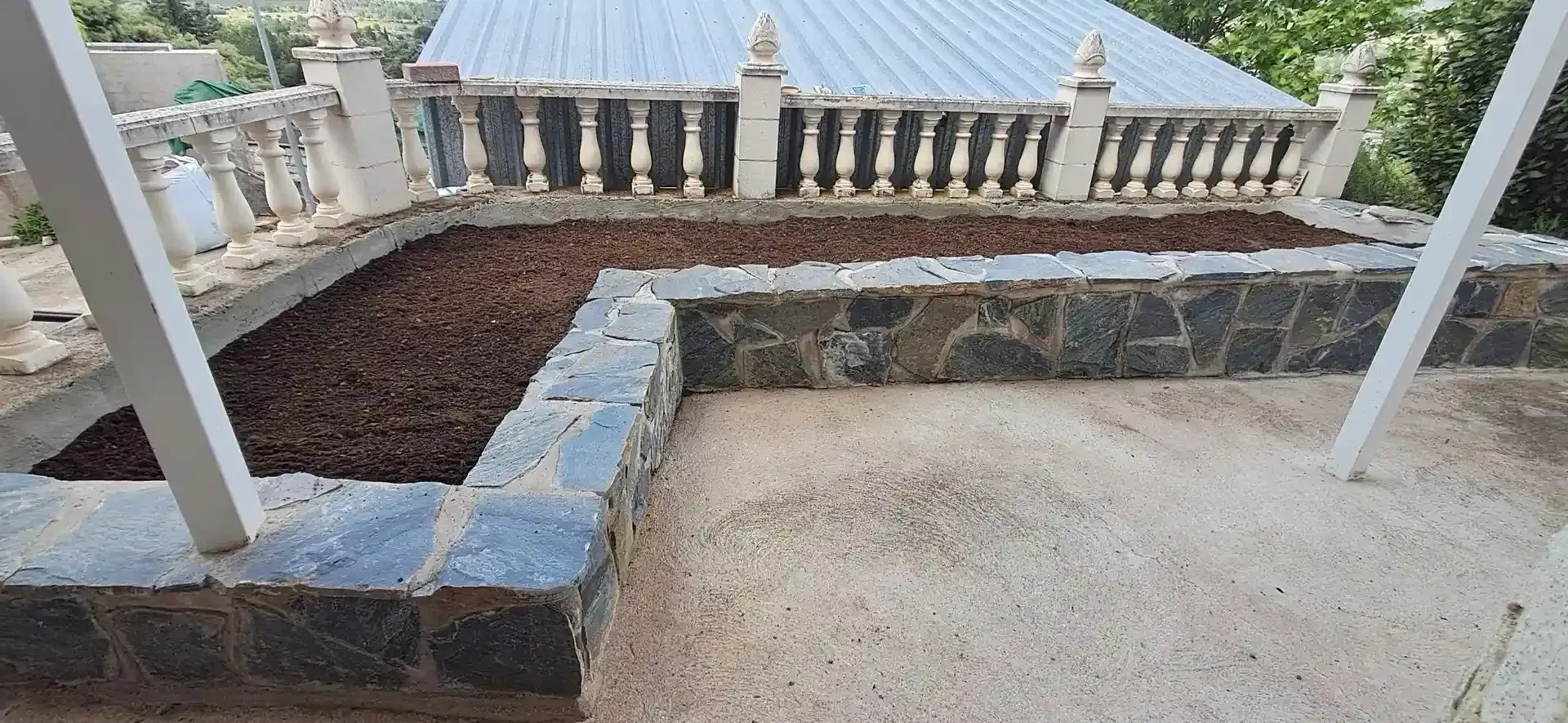 Un parterre elevado cubierto de mantillo está enmarcado por un muro de piedra. Al fondo se ven una barandilla blanca y un edificio.