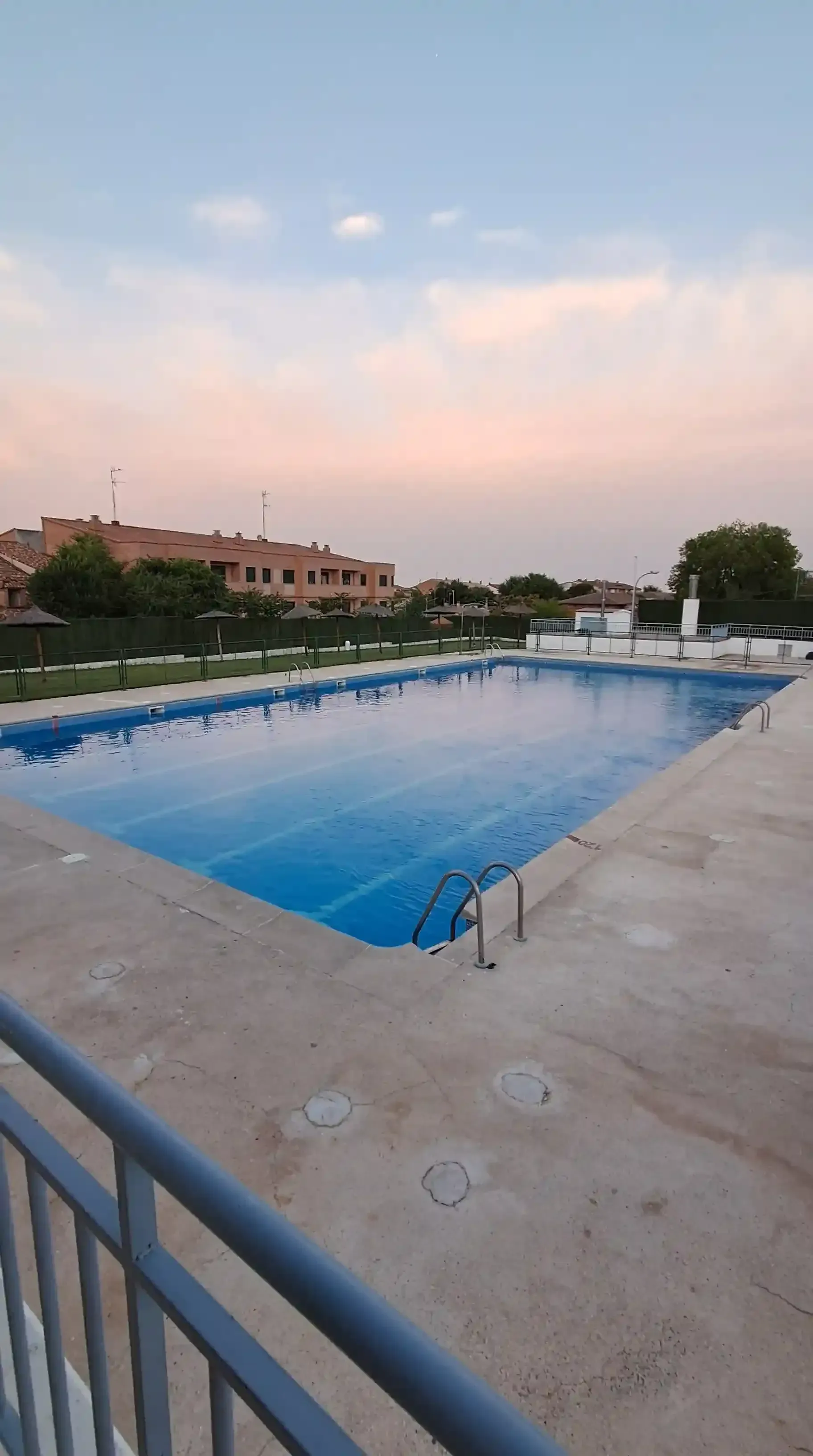 Piscina exterior vacía de agua azul, rodeada de hormigón. Edificios y un cielo colorido al fondo.