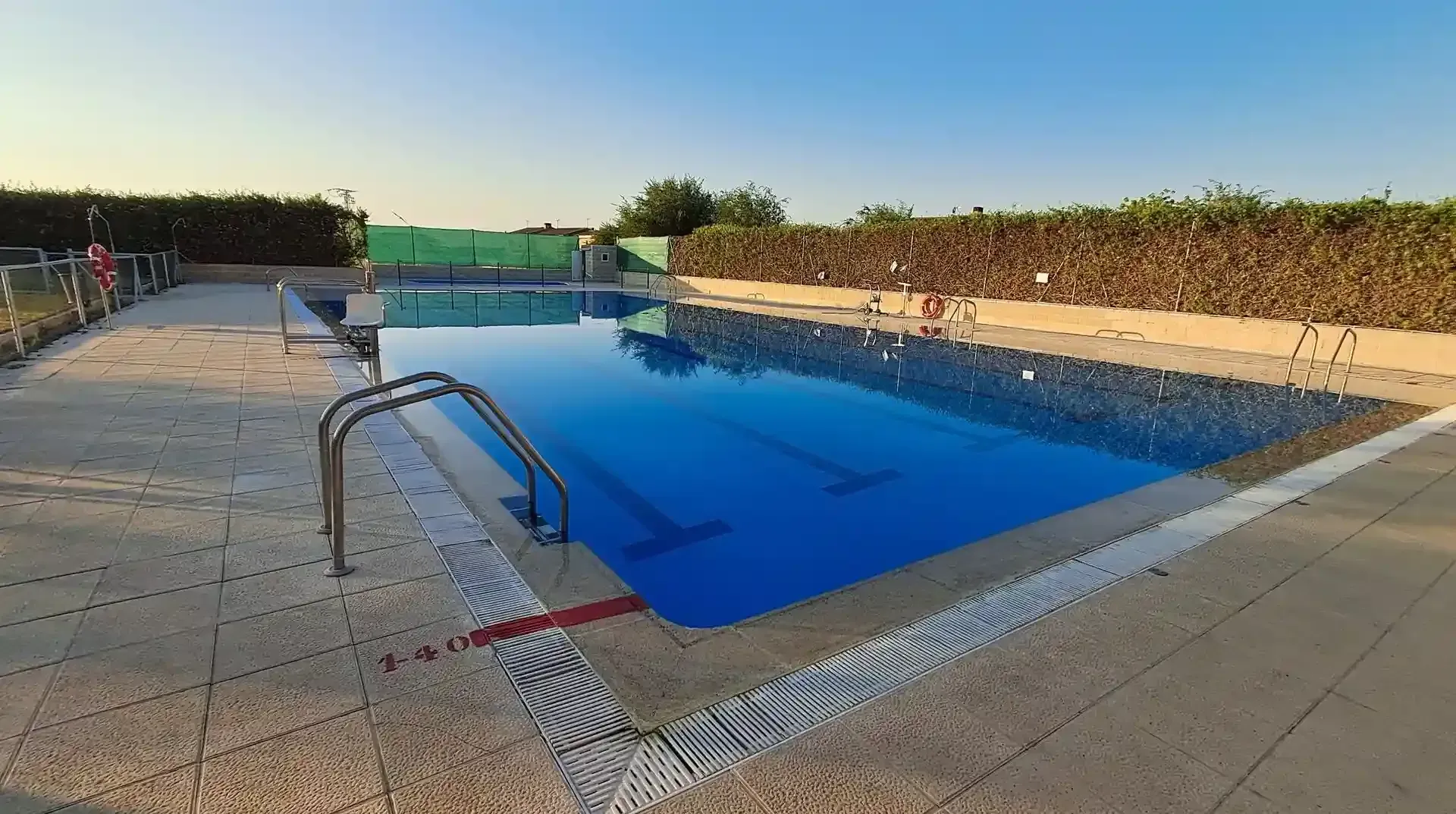 Piscina de agua azul, terraza de hormigón y seto verde bajo un cielo despejado.