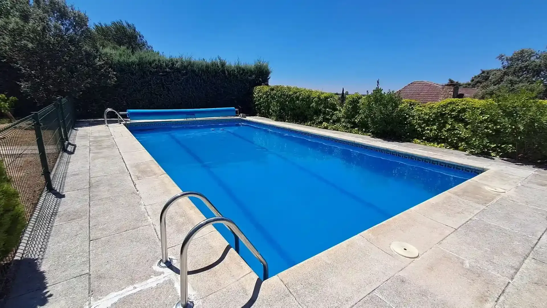 Piscina con cubierta azul, rodeada de patio de piedra, vegetación y una valla bajo un cielo despejado.