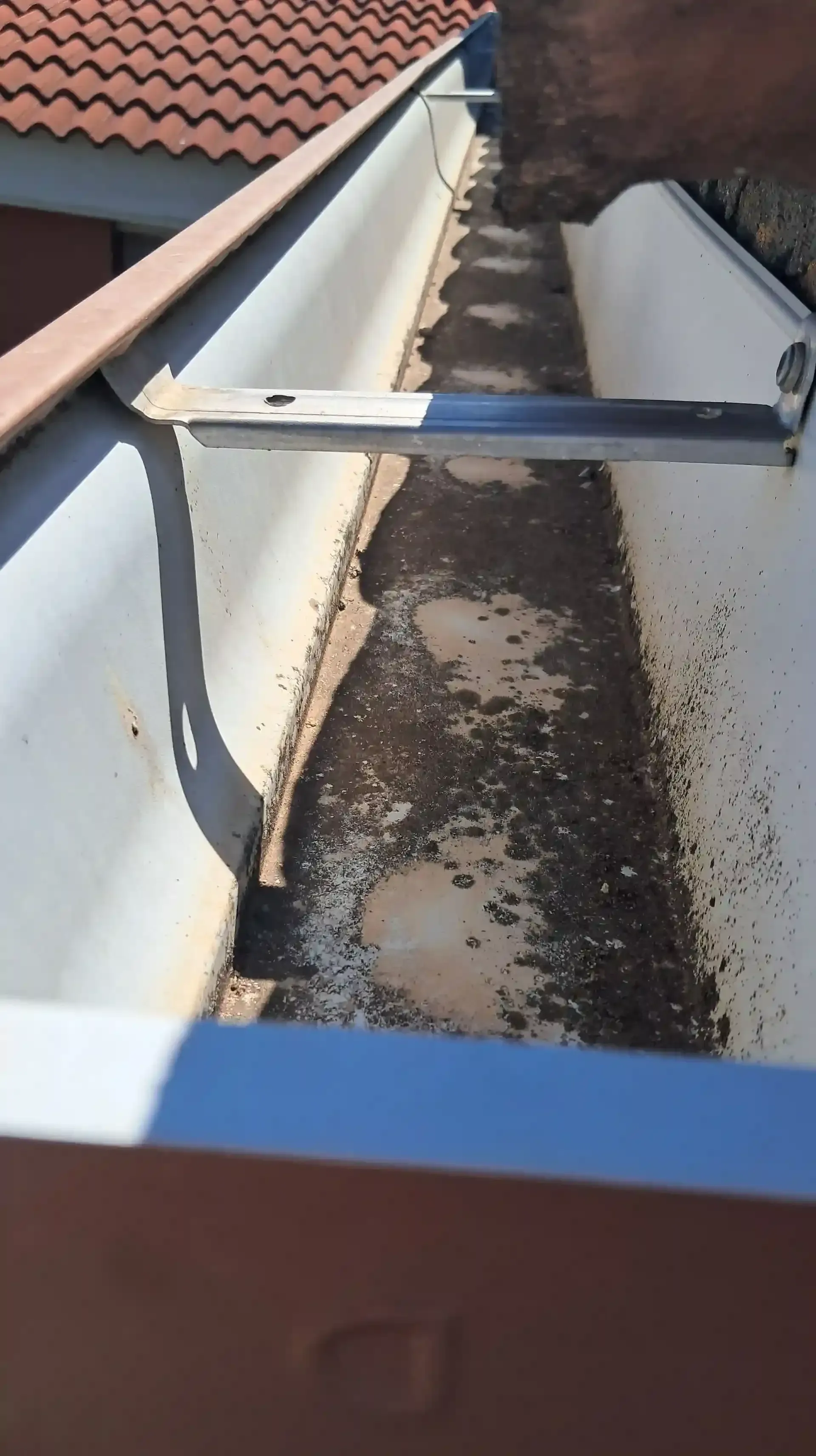 Canalón lleno de suciedad y escombros, visto desde arriba. Se aprecia un soporte metálico.