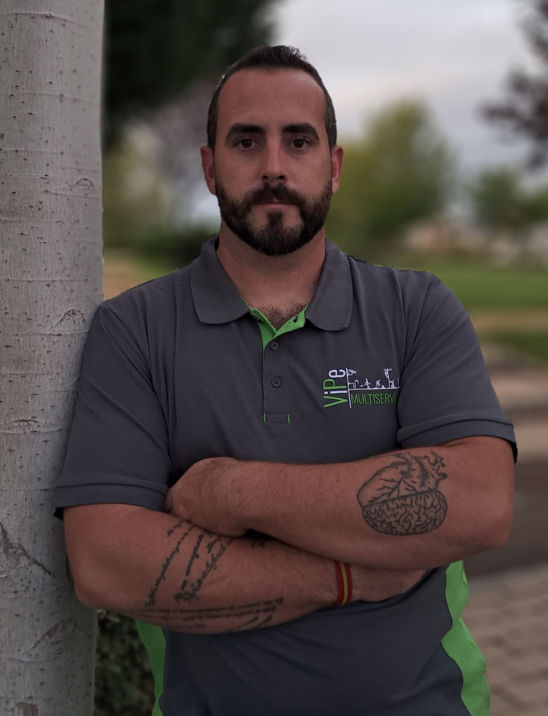 Hombre con camisa polo gris con los brazos cruzados apoyado en un árbol, el nombre 