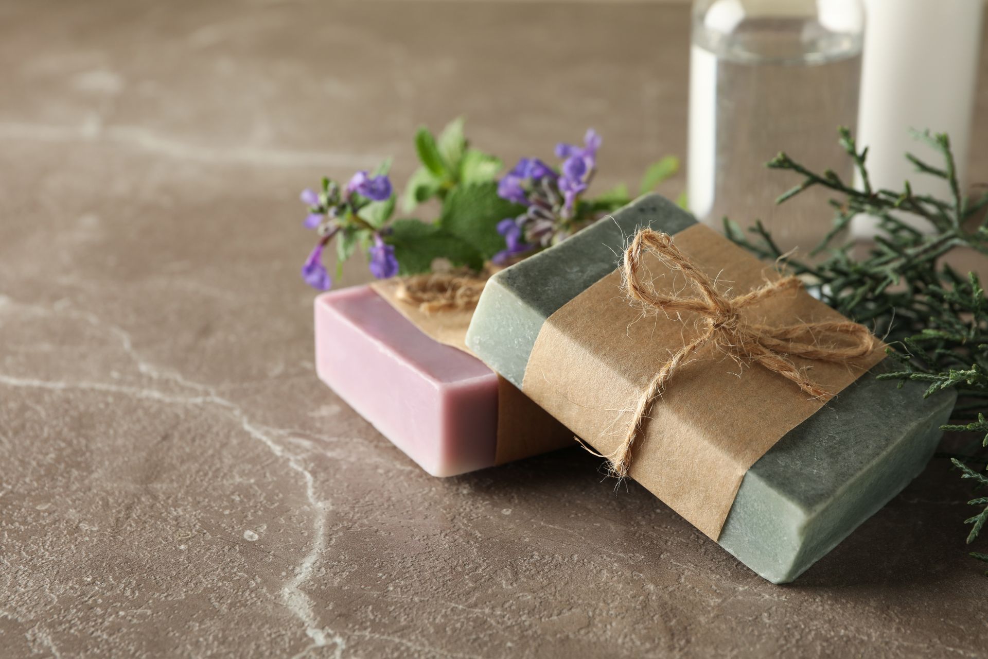 Zwei handgemachte Seifenstücke, in Papier mit Grünpflanzen auf einer strukturierten Oberfläche eingewickelt.