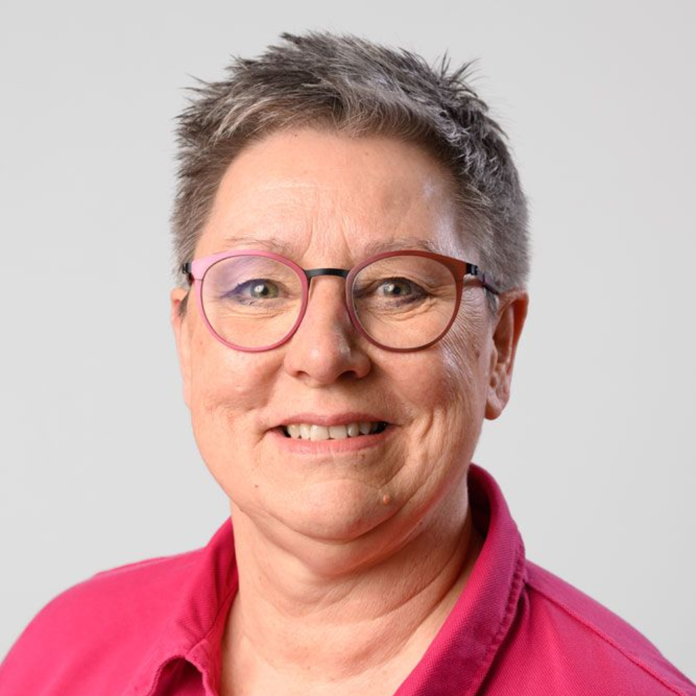 Eine lächelnde Person mit roter Brille sitzt in einem Auto und trägt ein rotes Hemd und einen grauen Kapuzenpullover.