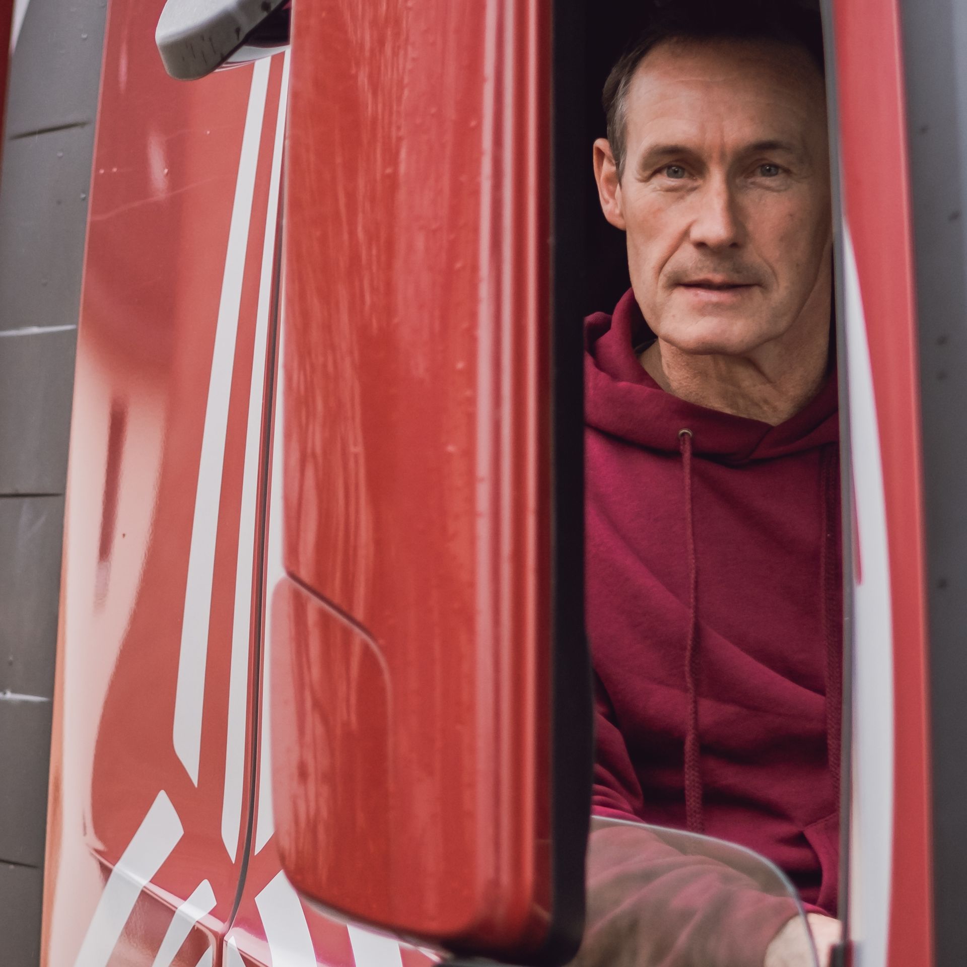 Ein Mann in einem roten Kapuzenpullover sitzt in der Tür eines roten Fahrzeugs und schaut in die Kamera.
