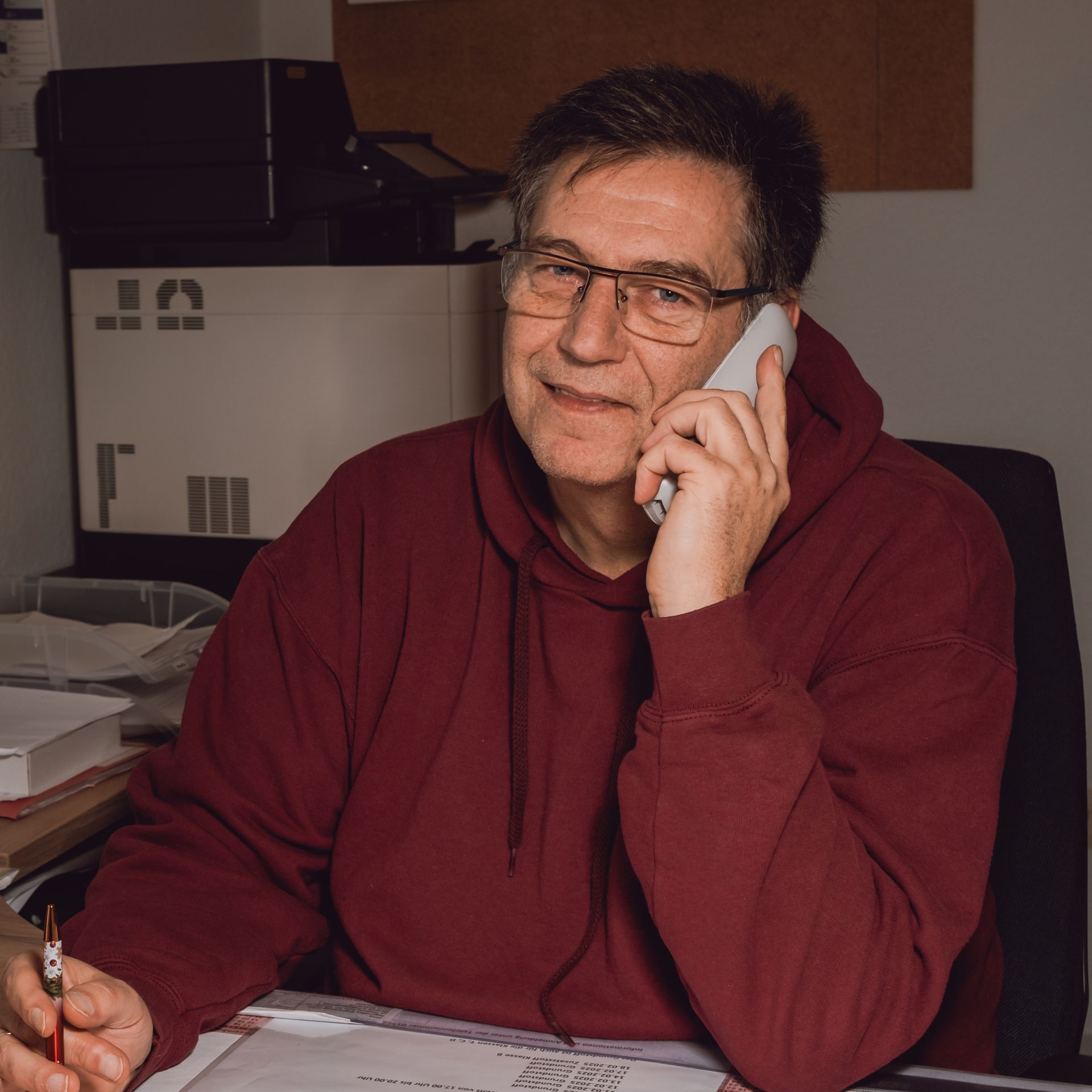Ein Mann in einem weinroten Kapuzenpulli und mit Brille sitzt in einem Büro an einem Schreibtisch und hält ein Telefon und einen Stift in der Hand.