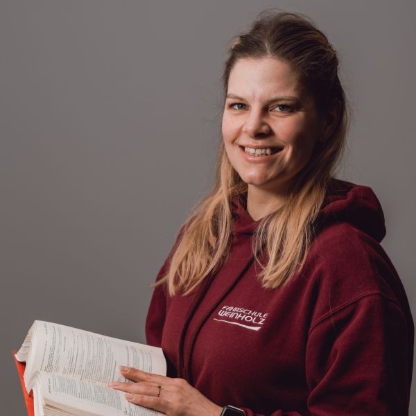 Frau in einem bordeauxroten Kapuzenpullover, die ein offenes Buch hält und vor einem grauen Hintergrund lächelt