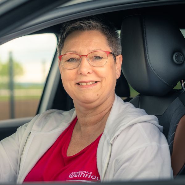 Eine lächelnde Person mit roter Brille sitzt in einem Auto und trägt ein rotes Hemd und einen grauen Kapuzenpullover.