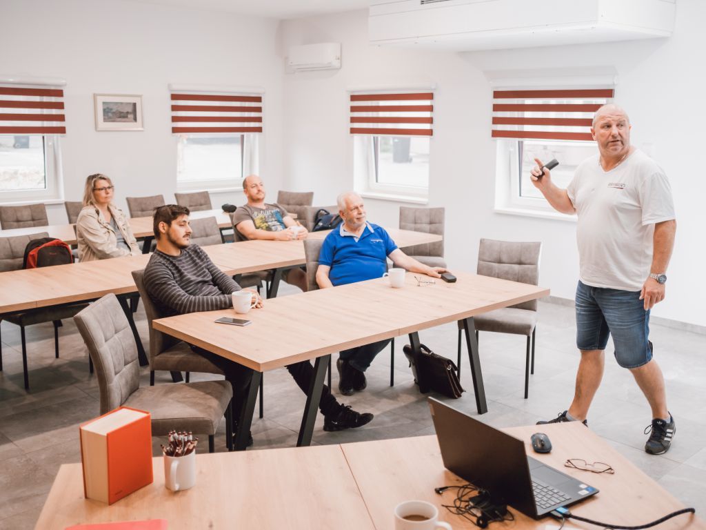 Präsentation im Besprechungsraum mit sitzenden Teilnehmern, die einem Mann zuhören, der neben einem Laptop und einem Projektor steht.
