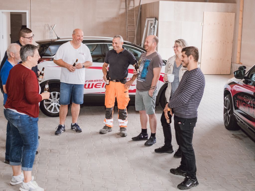 Eine Gruppe von Menschen unterhält sich in einer Garage neben einem weißen Sportwagen und einem roten Auto.