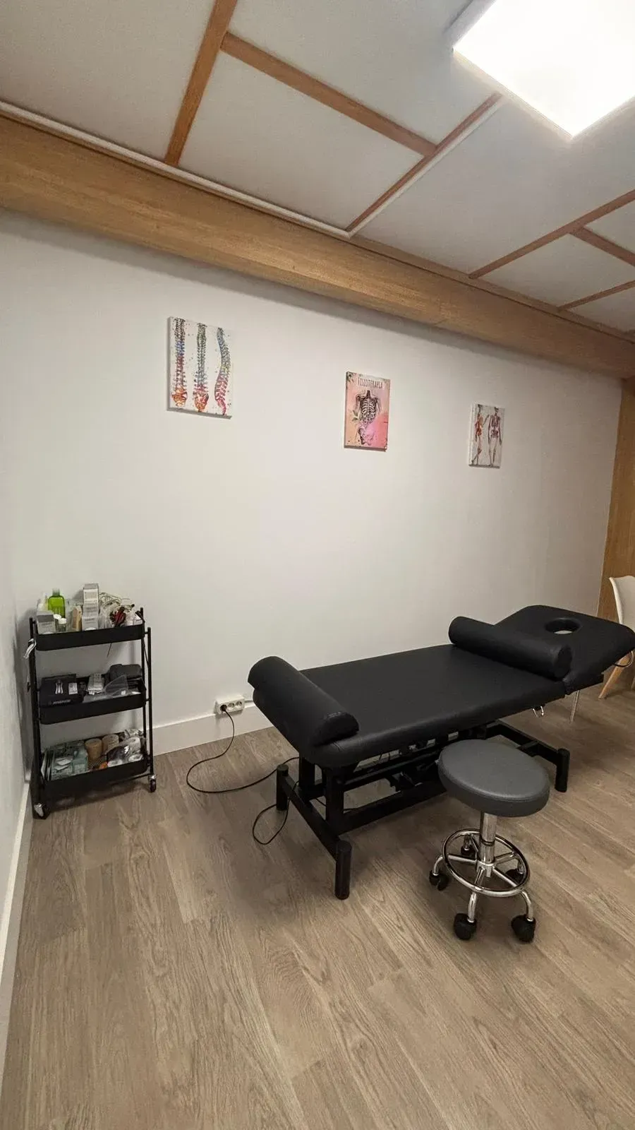Sala de tratamiento con mesa de masaje negra, taburete, pequeña estantería y arte de pared.