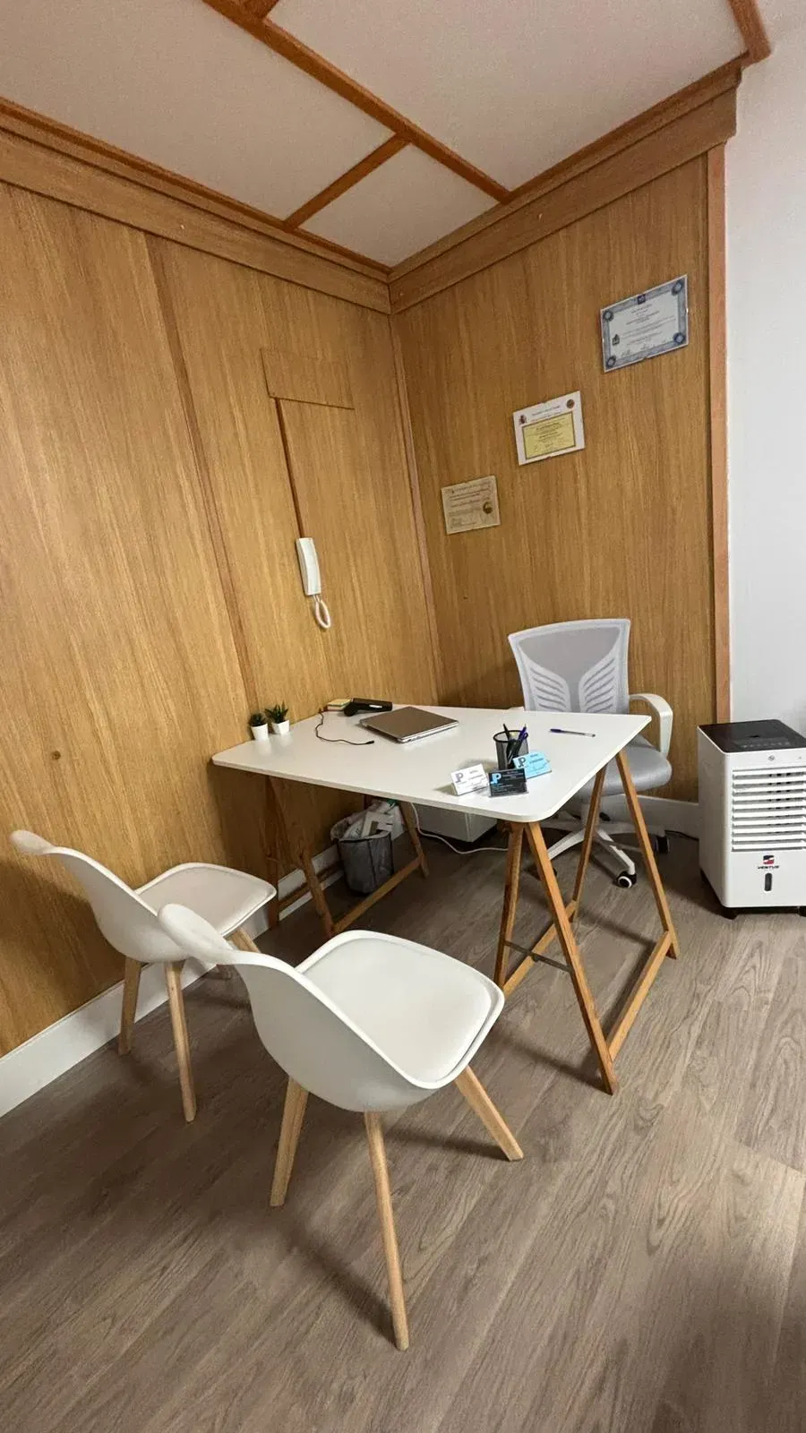 Una pequeña oficina con paneles de madera, un escritorio, dos sillas blancas y un archivador.