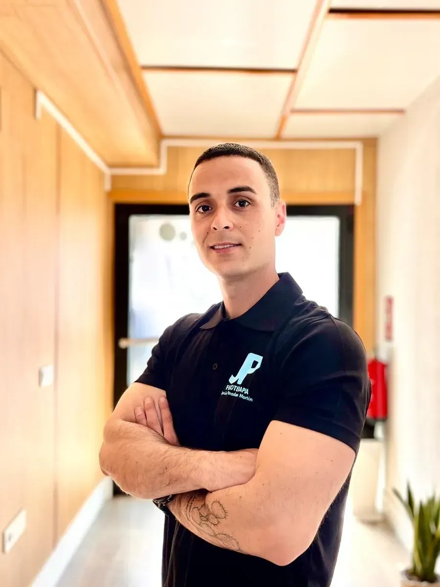 Hombre con los brazos cruzados, vestido con un polo negro, de pie en un pasillo.