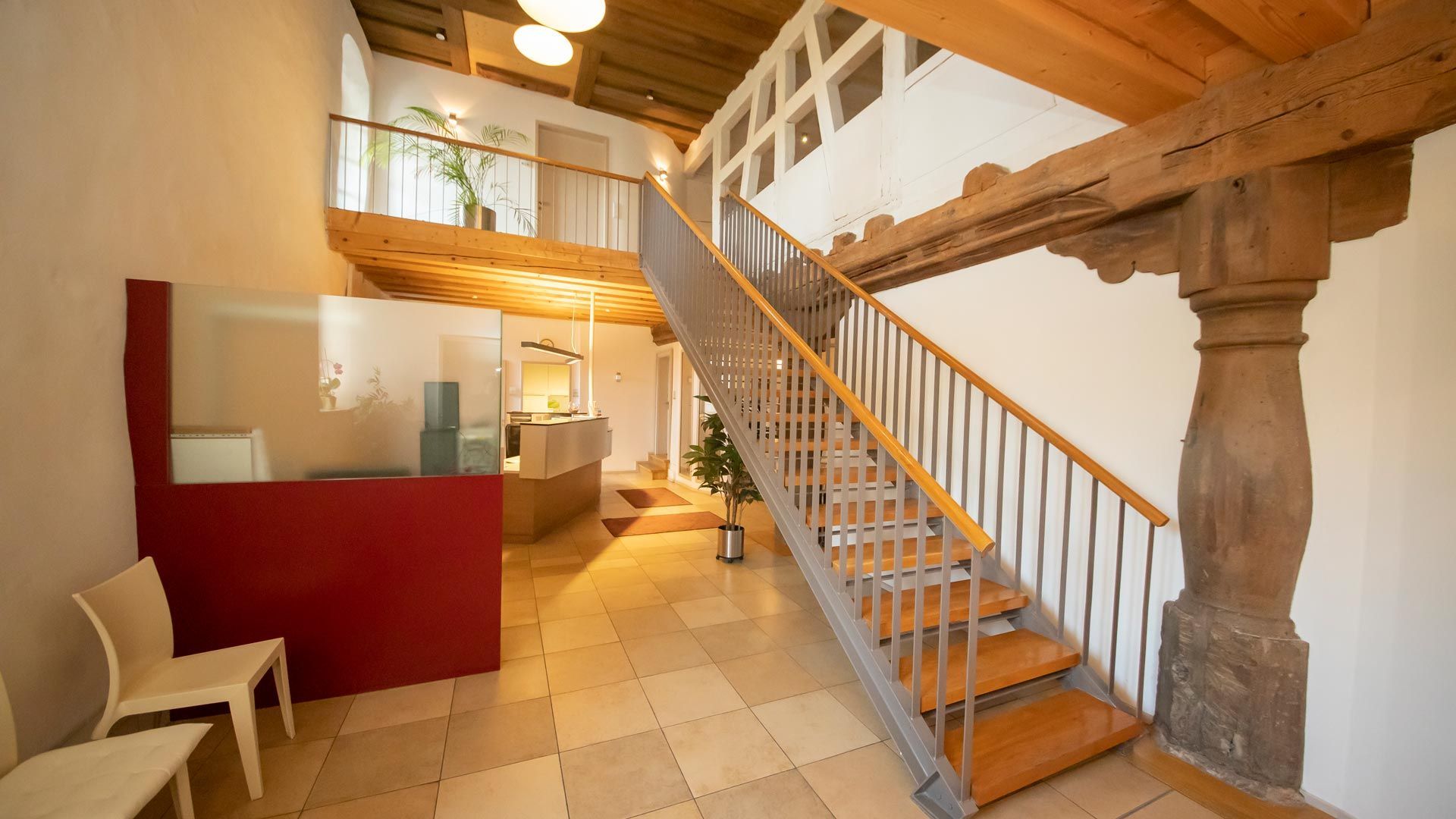 Rustikales Interieur mit Holztreppe, Steinsäule und rotem Empfangstresen unter warmem Licht