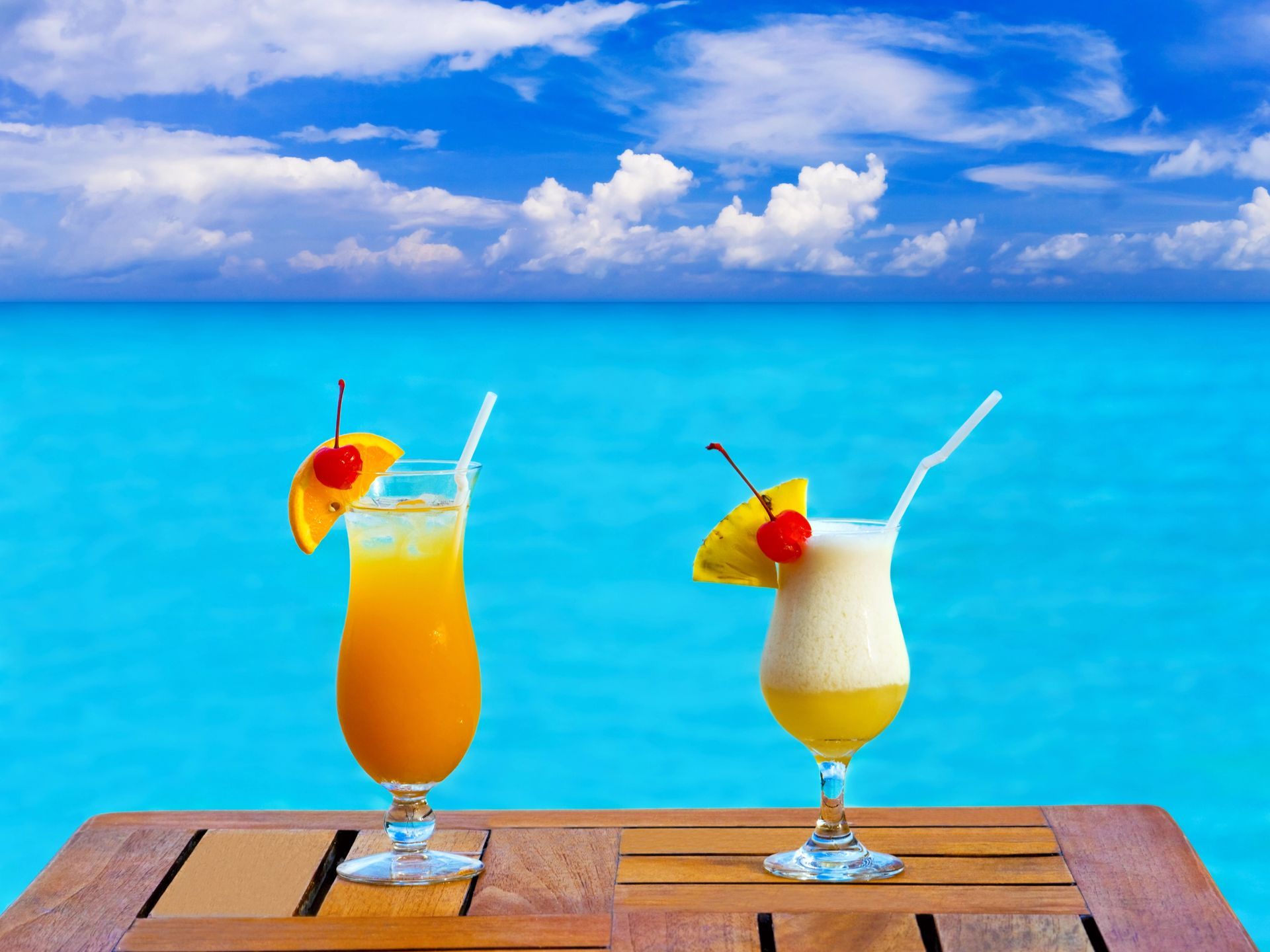 Two cocktails are sitting on a wooden table in front of the ocean.