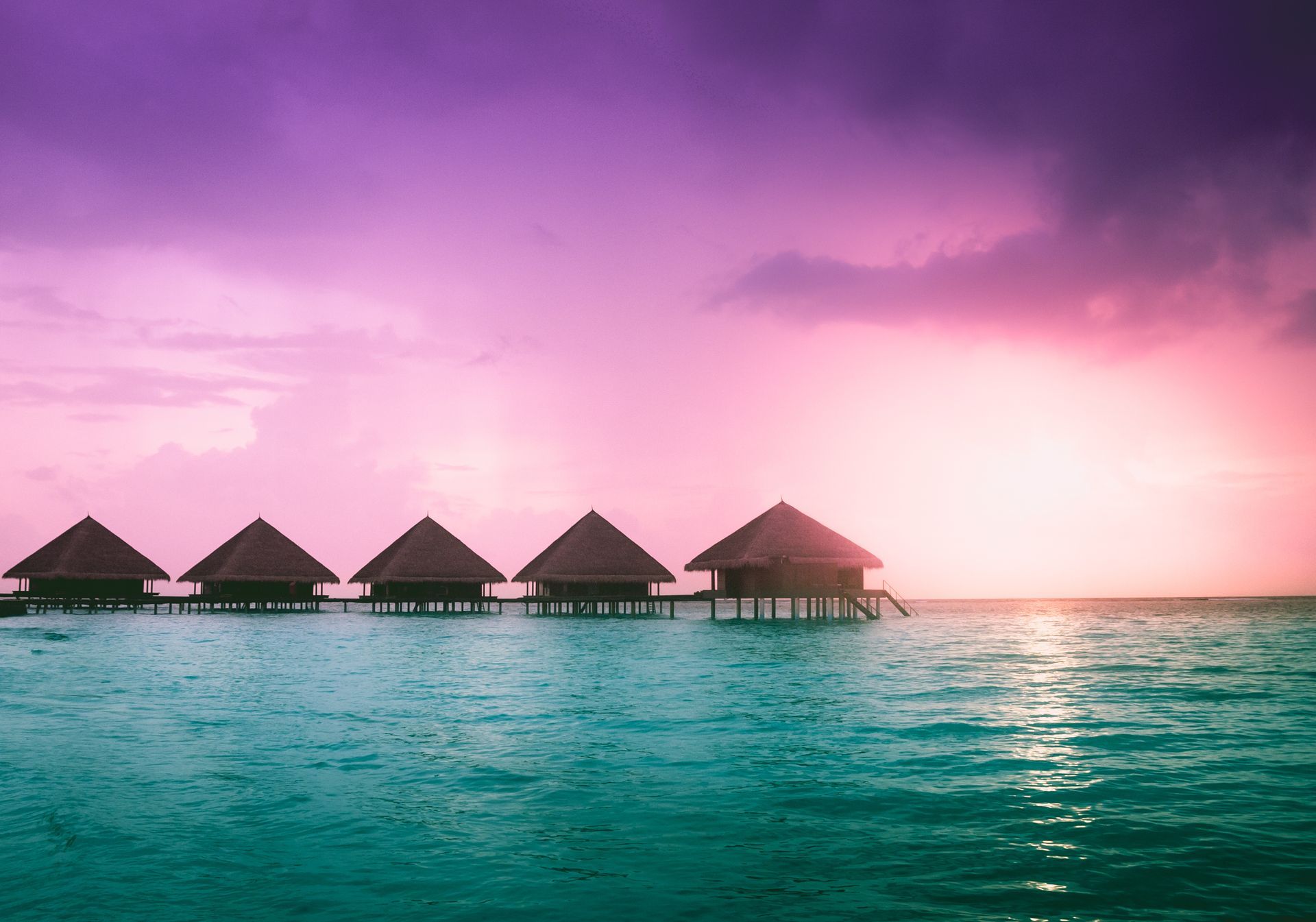 A row of bungalows on stilts in the middle of the ocean at sunset.