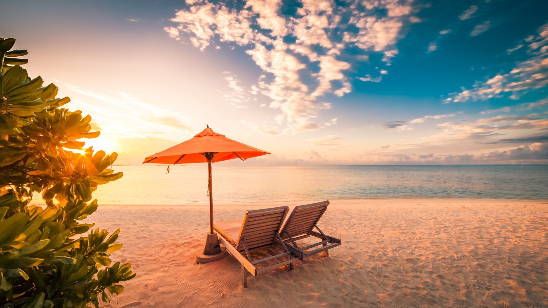 There are two chairs and an umbrella on the beach at sunset.