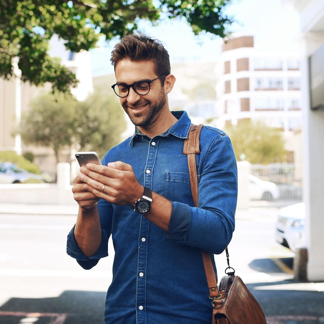 Un homme portant des lunettes et une chemise en jean sourit en utilisant son téléphone à l'extérieur, un sac à bandoulière est visible.