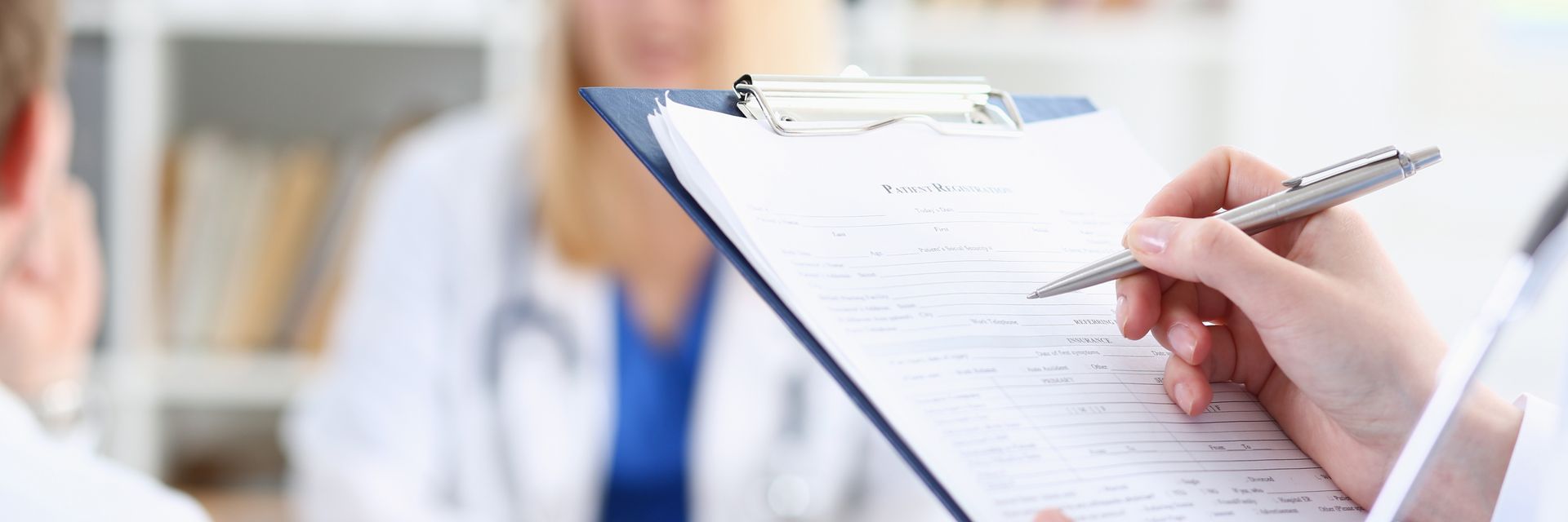 Un médecin prend des notes sur un bloc-notes pendant une consultation avec un autre médecin dans un cabinet médical à l'arrière-plan flou.