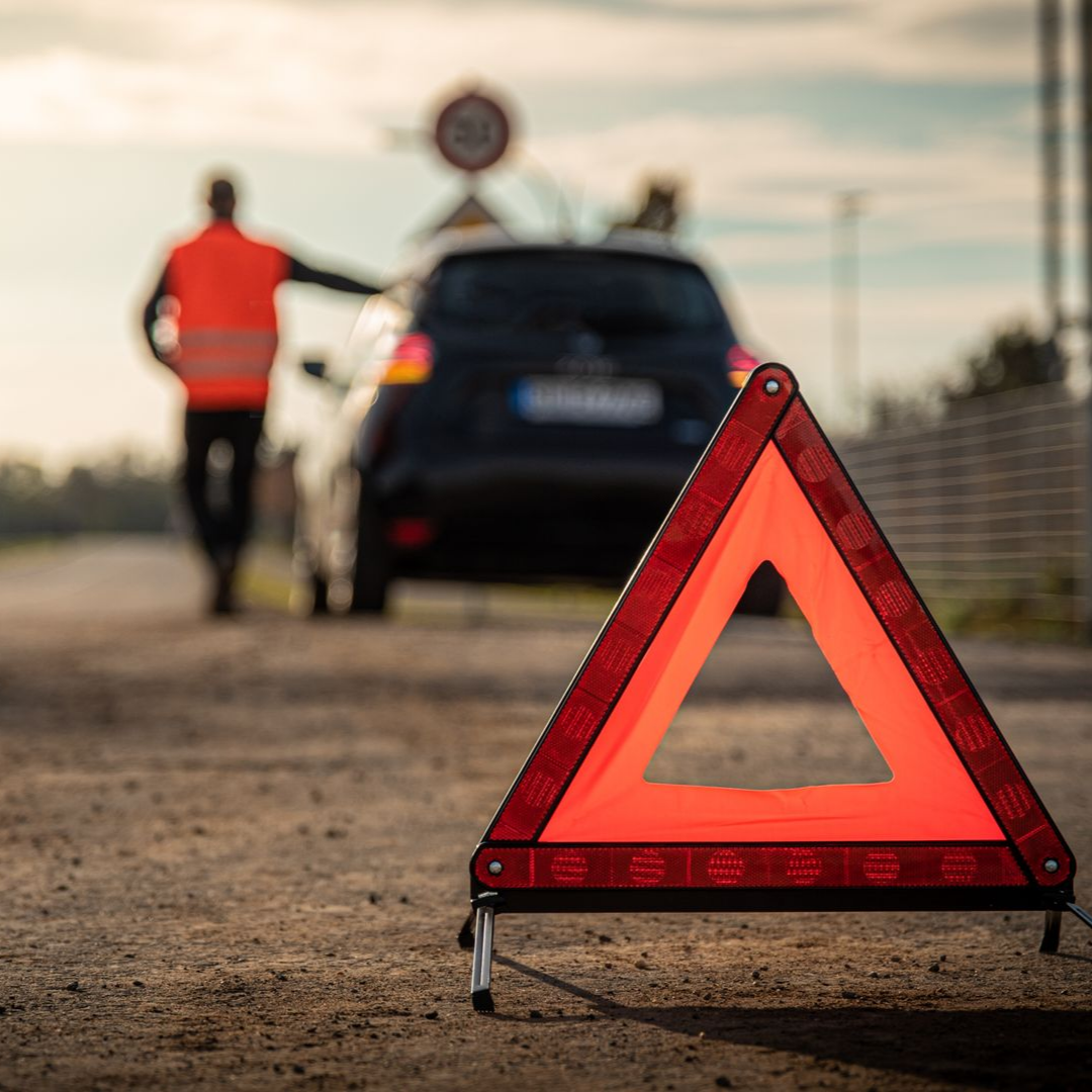 Un triangle de signalisation posé sur la route avec une voiture en panne en arrière-plan.