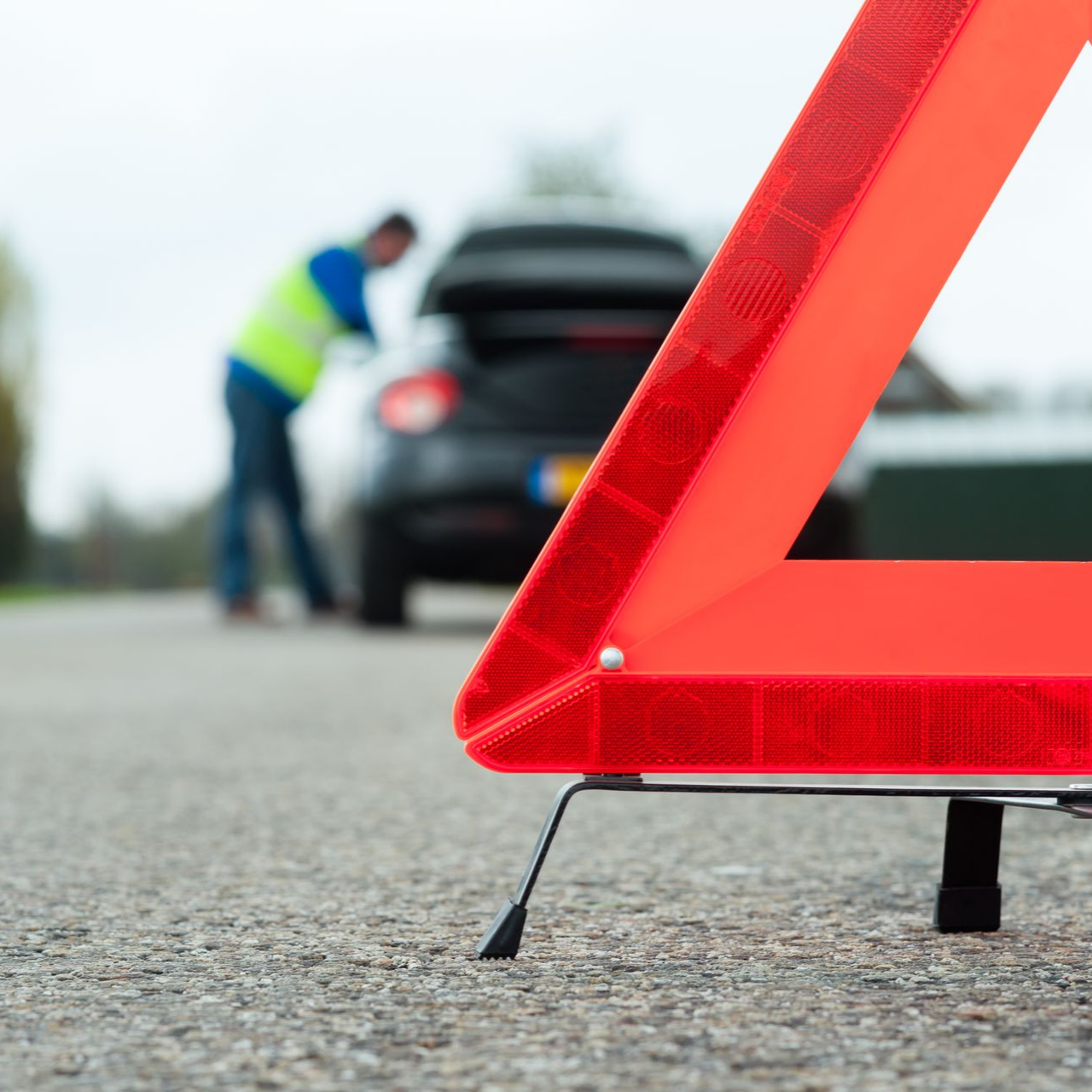 Triangle de signalisation rouge sur la route ; personne portant un gilet réfléchissant près d'une voiture.